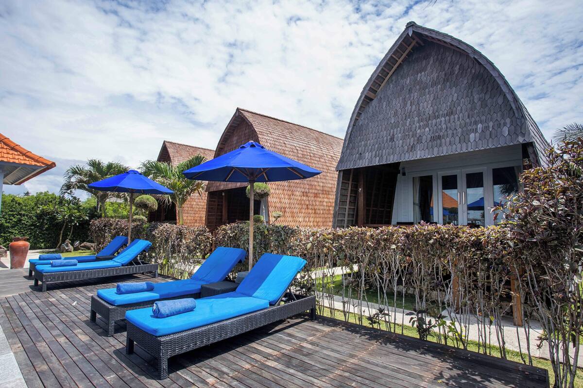 A resort with lounge chairs and umbrellas on a wooden deck, surrounded by well-maintained landscaping and a distinctive building with a curved roof.