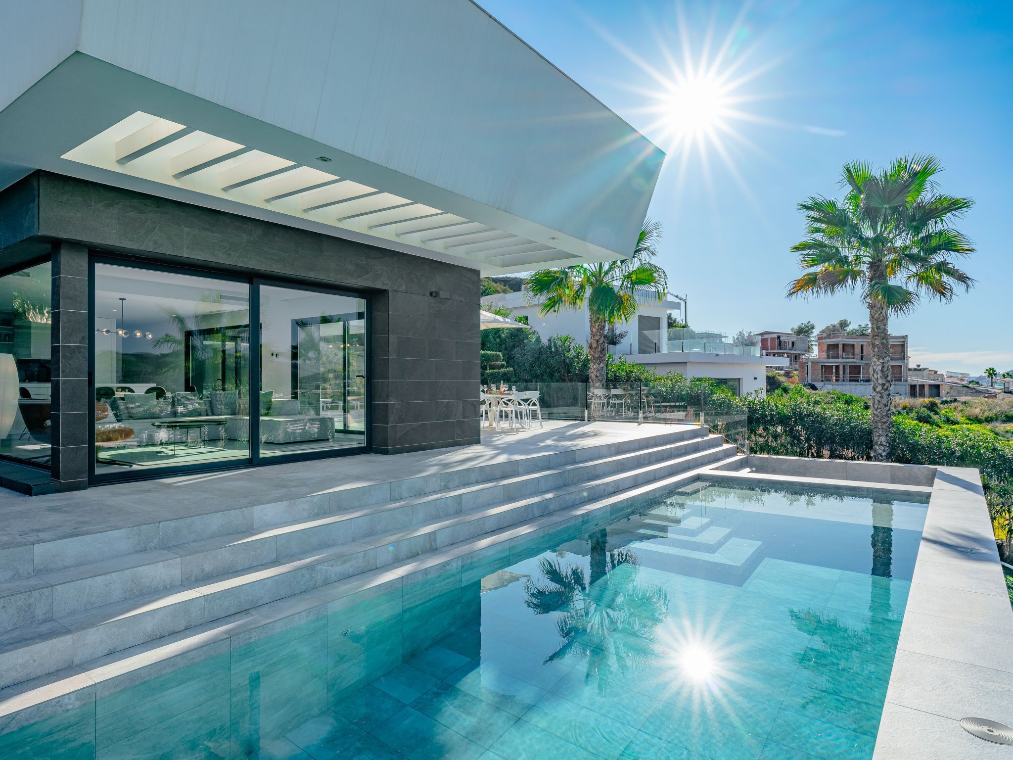 A modern house with a glass-enclosed patio, a swimming pool, and a view of palm trees and other houses.