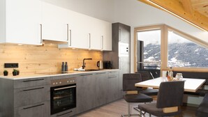Een moderne keuken met grijze kastjes, een houten aanrechtblad en een inbouwoven. Er is een zithoek met een tafel en stoelen, en een groot raam met uitzicht op een besneeerd landschap.