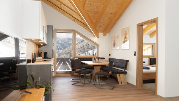 Een moderne keuken met houten balken, een eethoek met een bank en grote ramen met uitzicht op de omgeving.