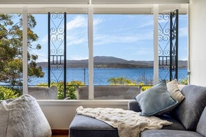 A room with a view of a lake and hills, a grey sofa with a blanket, and a window with metal bars. - Lemonade by the Beach: Serene Huon Valley Retreat with Water Views (Police Point)