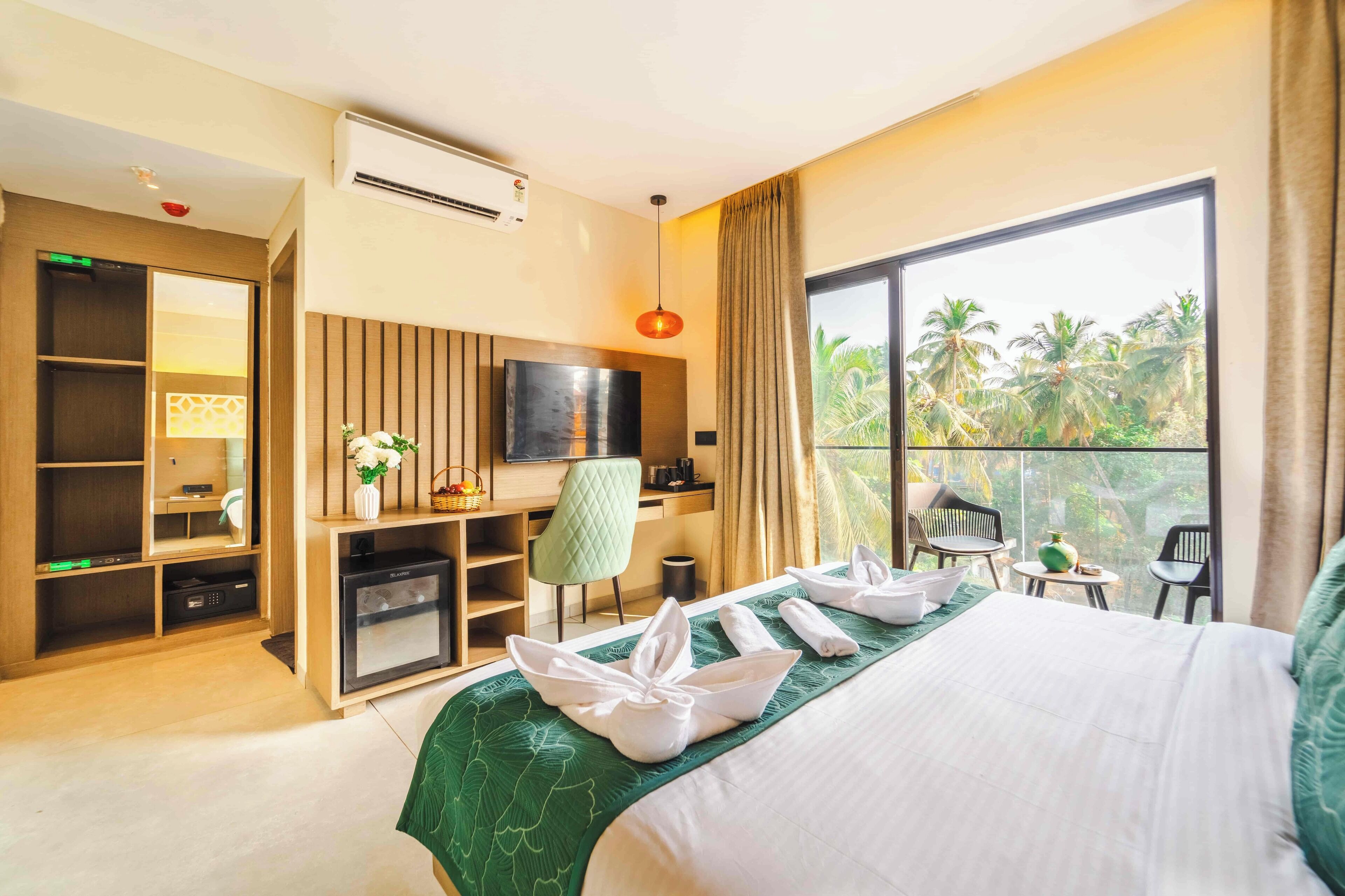 Habitación de hotel con cama, escritorio, televisión y balcón con vistas a palmeras.