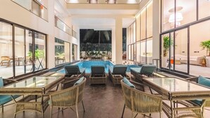 A spacious indoor pool area with wicker furniture and glass tables, surrounded by large windows and a view of the pool. - Vilmaris Luxury Hotel in Calangute (Calangute)