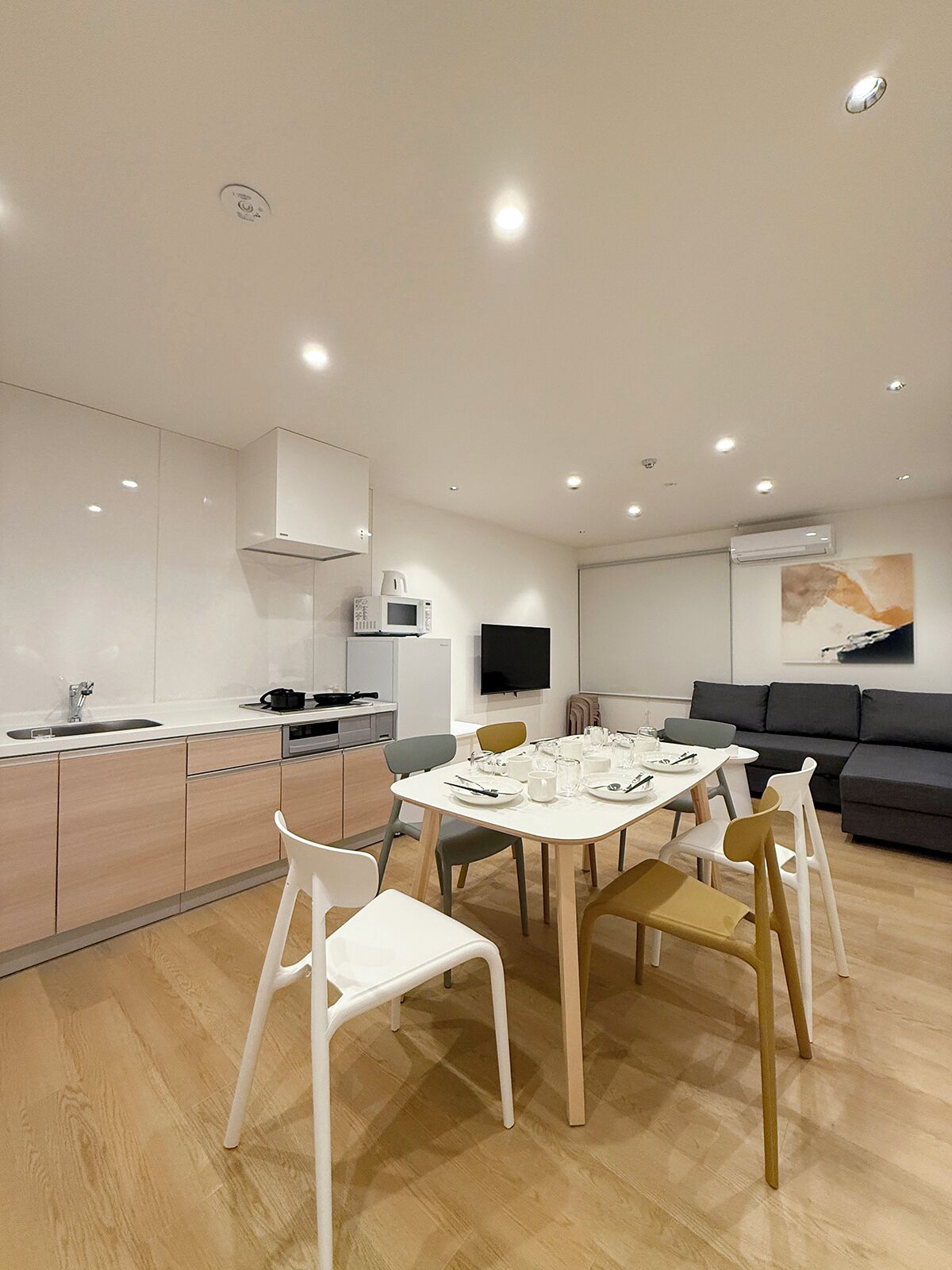 A modern kitchen with white cabinets, a dining table set for four, and a grey sofa in the living area.
