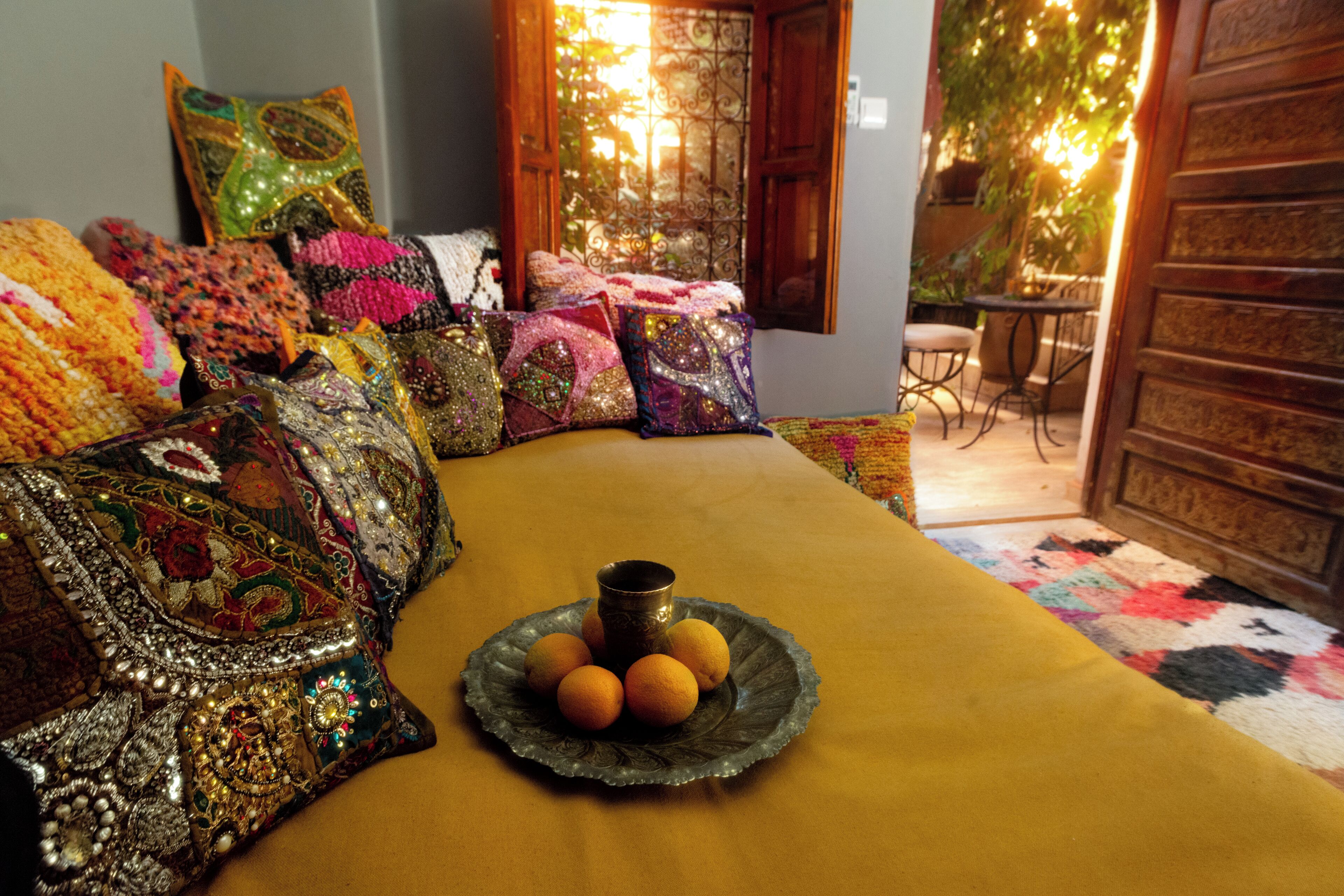 A room with a sofa adorned with colorful, patterned cushions, a small table with a decorative plate of oranges, and a view of an outdoor area with a wooden door and a table.