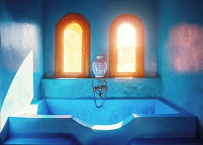 Une pièce avec une baignoire, deux fenêtres en arc de cercle et un vase bleu posé sur une étagère.
