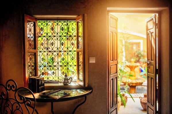 A room with a window featuring decorative iron bars, a table with books and a teapot, and an open door leading to an outdoor area with plants and a fountain. - Riad Roxanne (Marrakech)
