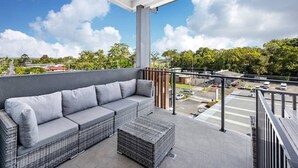 A balcony with a grey sofa and a small table, overlooking a parking area and greenery.