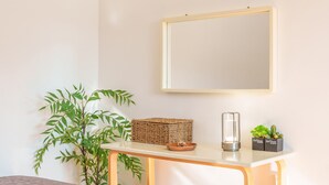 Une pièce avec un mur blanc, un miroir encadré, une table en bois, un panier en osier, un vase en verre et deux petites plantes en pot.