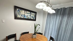 Un comedor con una mesa redonda de madera, seis sillas, una planta en maceta, un cuadro abstracto enmarcado y una lámpara en el techo.