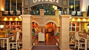 A hotel lobby with an arched entrance labeled "OSTERIA," featuring a central staircase, seating areas, and a view of the outdoor area. - Appartements im Freizeitpark Weissenhaeuser Strand (Wangels)