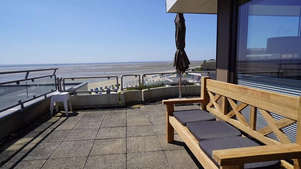 A balcony with a wooden bench, a small table, and a glass railing overlooking a beach.
