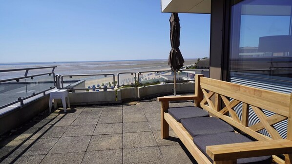 A balcony with a wooden bench, a glass railing, and a view of the beach.