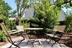 Un área de estar al aire libre con dos sillas de madera y una mesa, rodeada de vegetación y un edificio con techo de tejas.