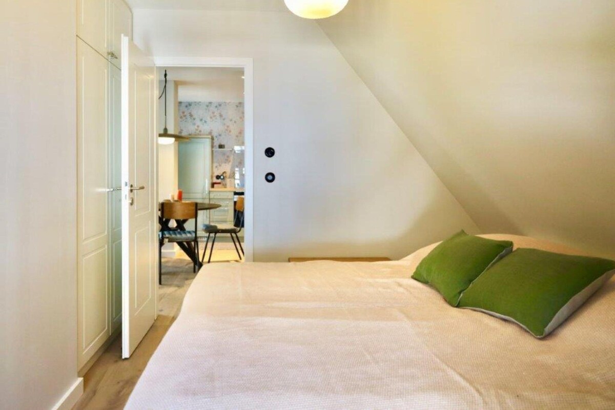 Un dormitorio con cama, almohada verde, escritorio con silla y una cocina al fondo.