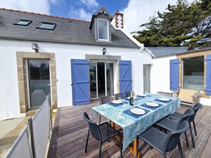 An outdoor patio with a table set for dining, surrounded by chairs, and a wooden deck. - Charming Penty 2-4 People, Morlaix bay (Plougasnou)