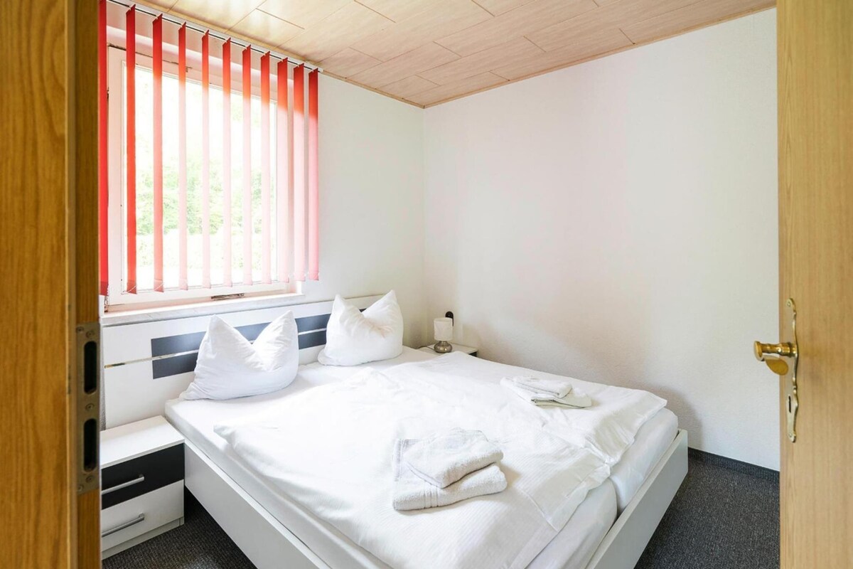 Ein Schlafzimmer mit Bett, Kissen, Nachttisch, Lampe und einem Fenster mit Jalousien.