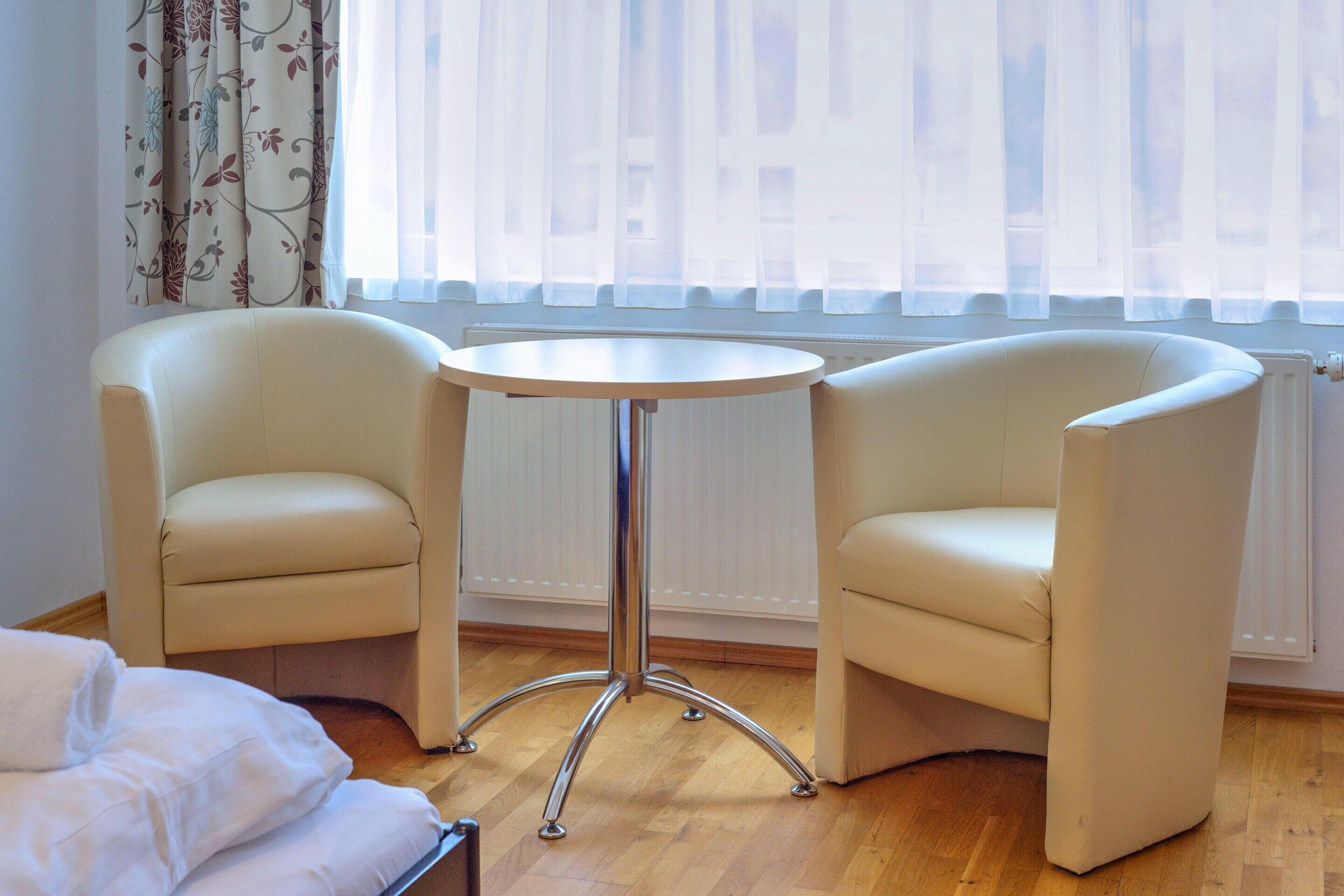 Een kamer met twee witte fauteuils, een kleine ronde tafel en een bed.