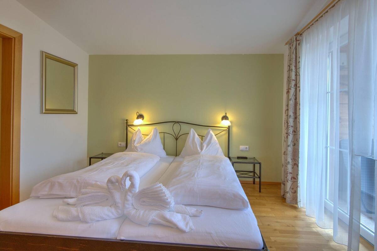 A neatly made bed with white linens and towels arranged in a swan shape, a bedside table with a lamp, and a window with sheer curtains.