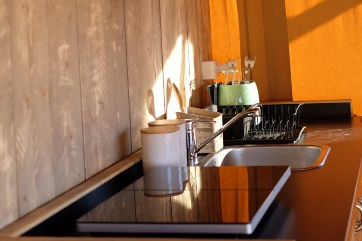 Un interior de madera con área de cocina, que incluye un lavabo, estantes con frascos y un ventanal que deja entrar luz natural.