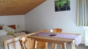Ein Zimmer mit einem Bett, einem Tisch und einem Stuhl. Ein Fenster mit Vorhängen und ein Bild an der Wand.