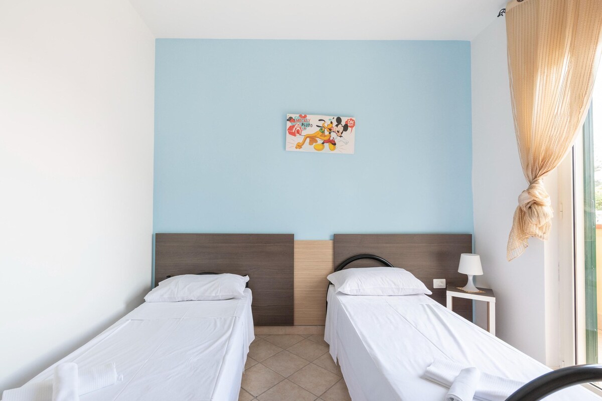 Quarto de hotel com duas camas de solteiro, cabeceira, mesa de cabeceira pequena com abajur e janela com cortina.