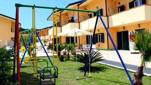 Un terrain de jeux avec des balançoires et un toboggan, entouré d’un bâtiment doté de balcons et d’un espace extérieur aménagé pour s’asseoir.