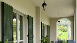 A covered patio area with outdoor seating, green shutters, and a view of the garden. - A Casa Guesthouse (Mombaruzzo)