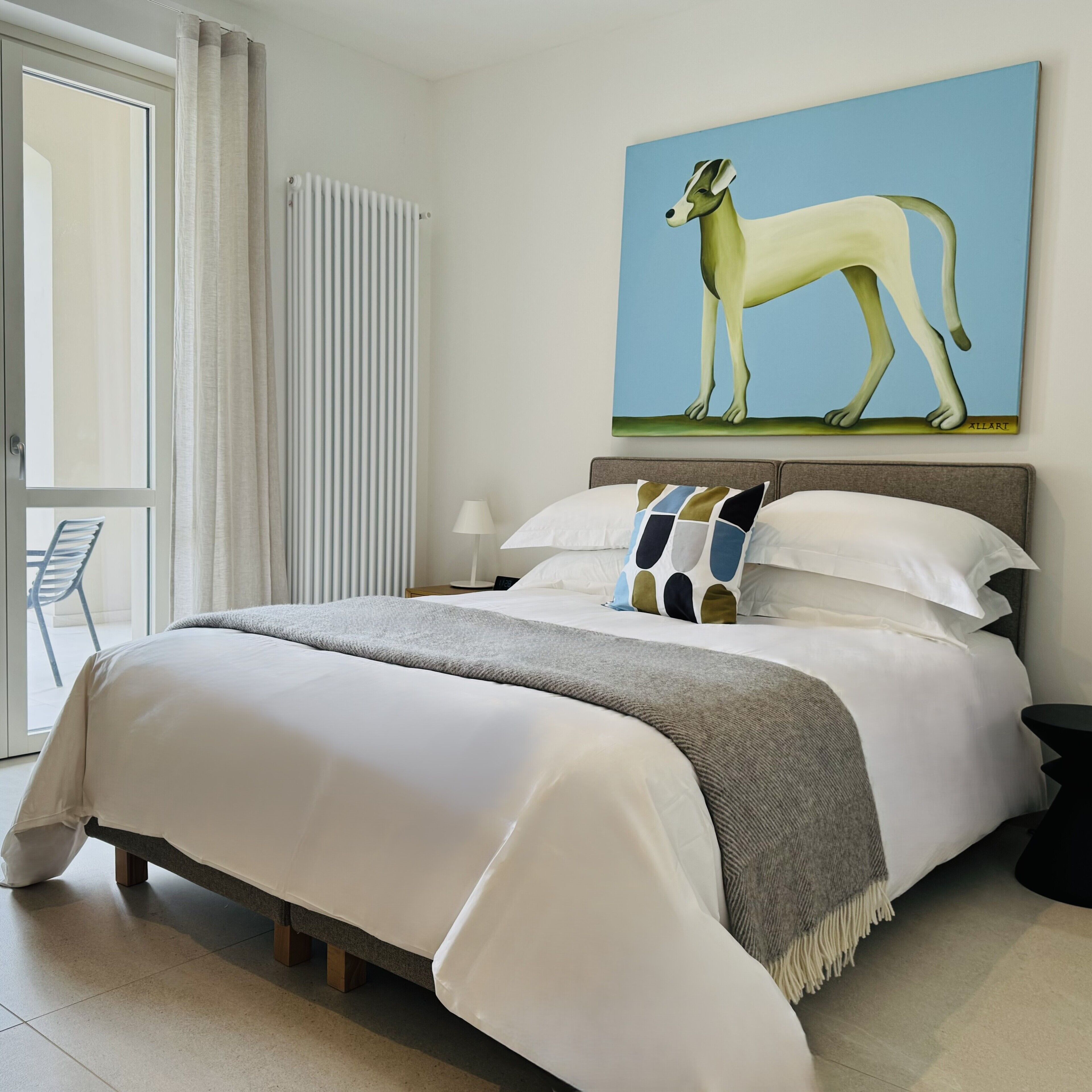 A bedroom with a large bed, a painting of a dog, a window with curtains, and a chair by the window.