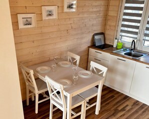 Une pièce aux murs lambrés, avec une table à manger dressée pour quatre personnes, une petite cuisine équipée d’un évier et d’une cuisinière, et des tableaux encadrés accrochés au mur.