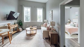 A spacious living room with a ceiling fan, a flat-screen TV mounted on the wall, a wooden coffee table, a beige sofa, and a small side table with a lamp. - Sanctuary Co Loft in Downtown Johnson City (Johnson City)