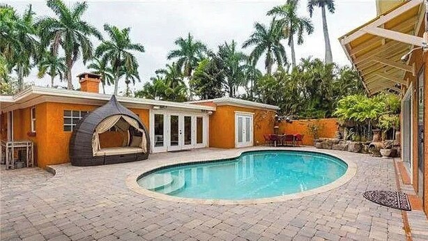 Un área de piscina con una piscina curva, un espacio de estar techado y un edificio con techo de azulejos.