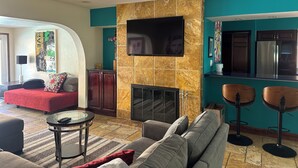 Una sala de estar con chimenea, un sofá, un televisor montado en la pared y una zona de cocina con refrigerador.