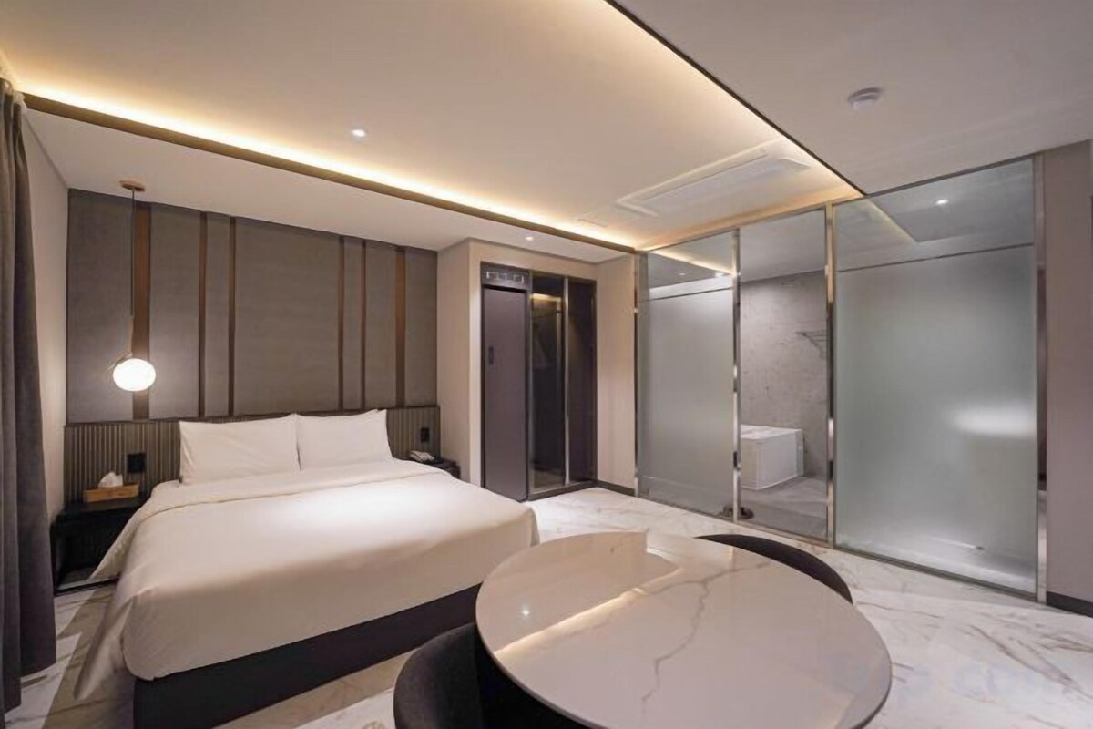 A modern hotel room with a large bed, a round marble table, a separate bathroom, and a glass-enclosed shower area.