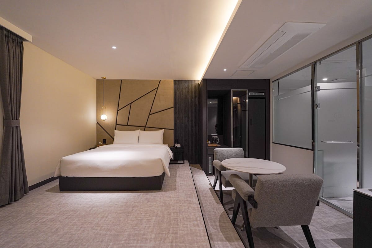A modern hotel room with a large bed, a small table with two chairs, and a glass partition wall.