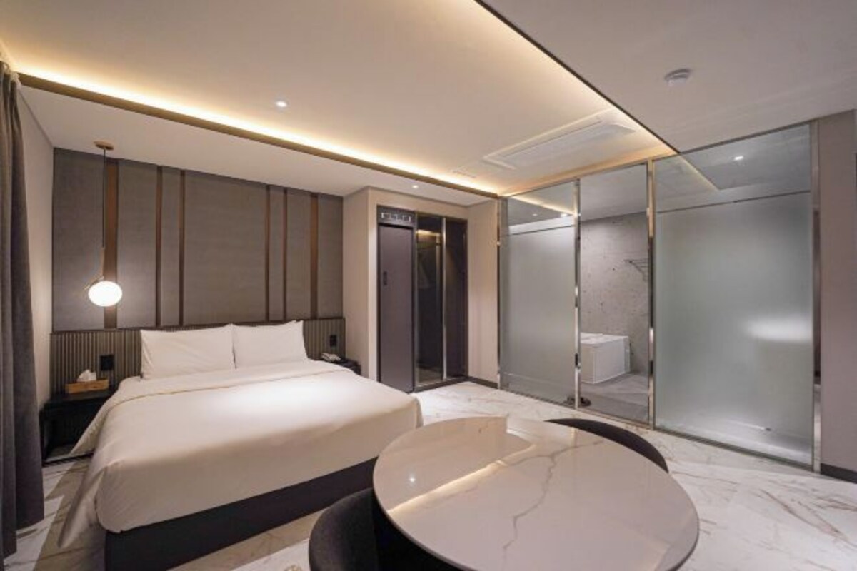 A modern hotel room with a large bed, a round table, a glass-enclosed shower, and a separate bathroom.