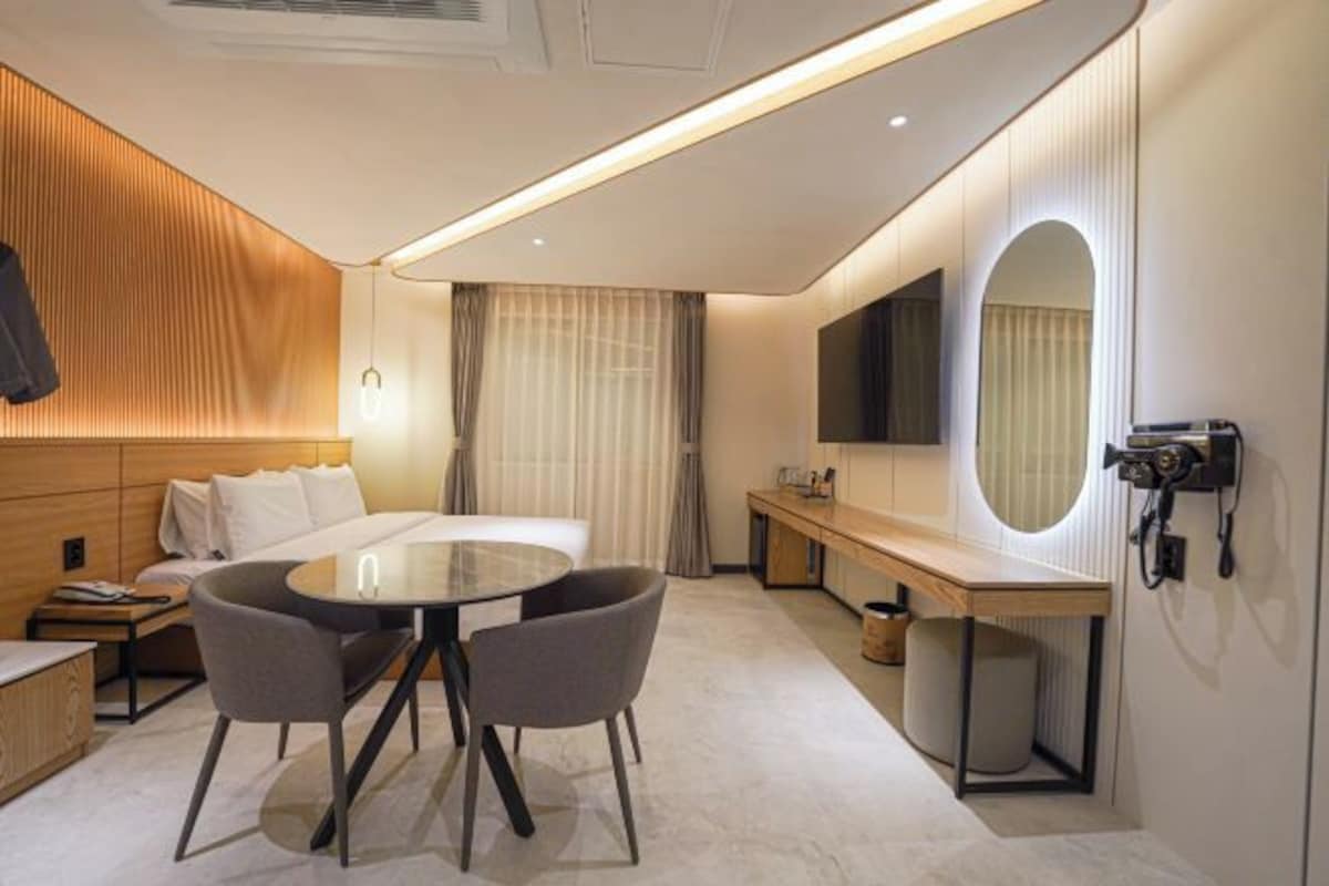 A modern hotel room with a round dining table, two chairs, a sofa, a large mirror, a TV, and a minibar.
