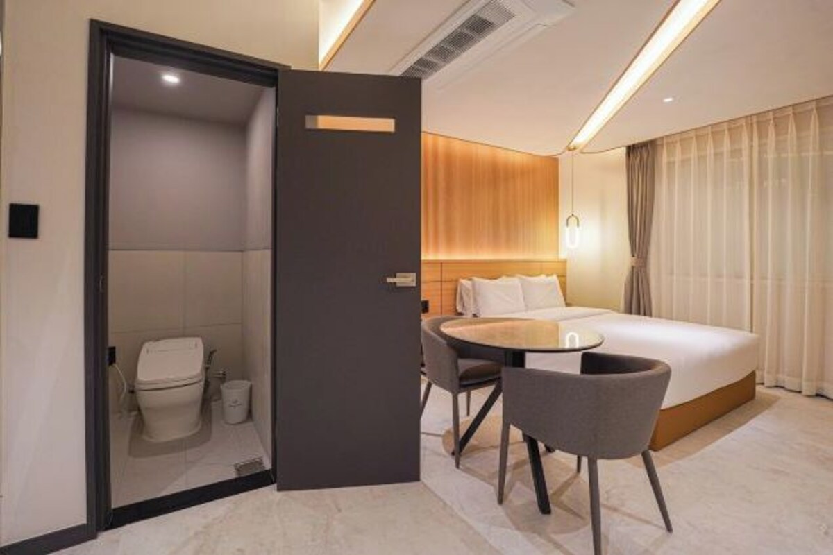 A hotel room with a bed, a small table, a chair, a toilet, and a door leading to a shower area.