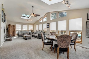 A spacious living room with a high ceiling, large windows, a ceiling fan, and a dining area with patterned chairs.