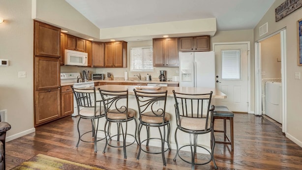 A kitchen with wooden cabinets, a central island, and a dining area with metal chairs. - Crestview Place! 2 Mins to UCCS, Patio, and BBQ (Colorado Springs)