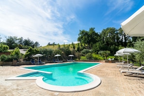 A rectangular swimming pool with a tiled edge, surrounded by lounge chairs and umbrellas, set against a backdrop of trees and a clear sky.