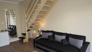 Ein Wohnzimmer mit einer Holztreppe, einem schwarzen Sofa mit grauen Kissen, einem kleinen Holzschemel, einem weißen Schrank und einem Spiegel, der den Raum widerspiegelt.