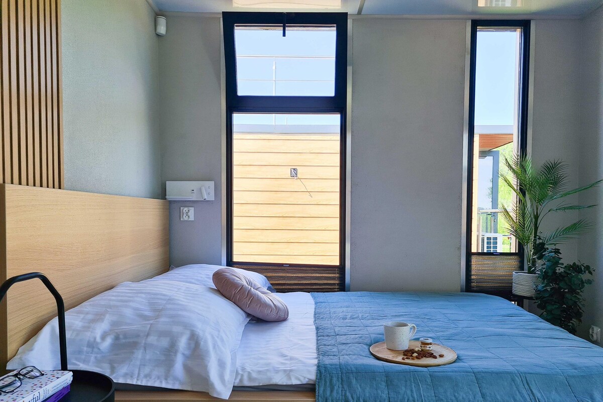 Een moderne slaapkamer met een groot bed, een nachtkastje met een boek, een kopje en een bordje noten, en een raam met uitzicht.