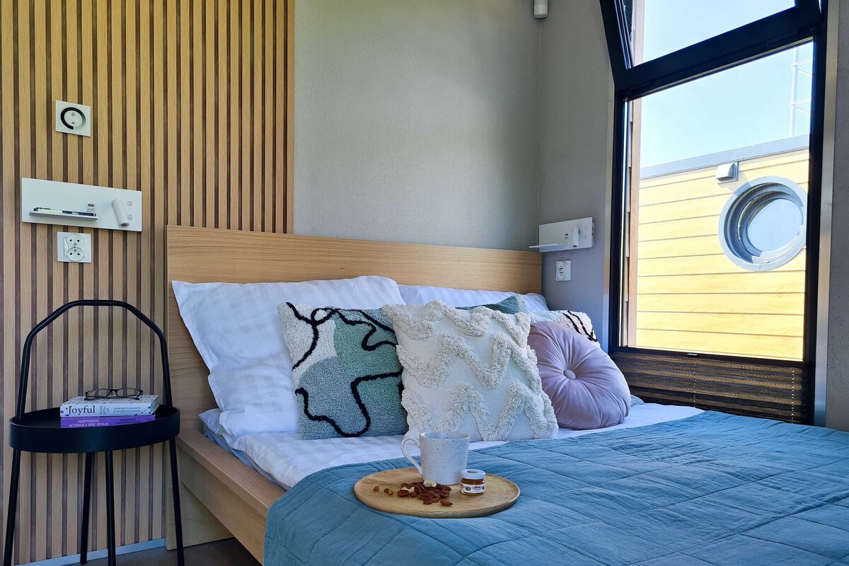 Een keurig opgemaakt bed met een blauwe sprei, een nachtkastje met boeken, en een raam met uitzicht op een gebouw.