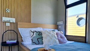 Een net opgemaakt bed met een blauwe sprei, een nachtkastje met boeken en een raam met uitzicht op een gebouw.