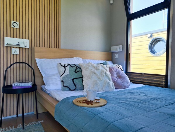 Ein ordentlich bezogenes Bett mit einer blauen Stegdecke, ein Nachttisch mit Büchern und ein Fenster mit Blick auf ein Gebäude.