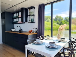 Een moderne keuken met een centraal eiland, een eettafel met stoelen en uitzicht op groen door grote ramen.