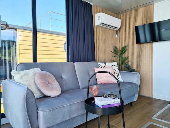 Un salon moderne avec un canapé gris, une petite table d’appoint noire, un climatiseur fixé au mur, un téléviseur et une grande fenêtre avec des rideaux.