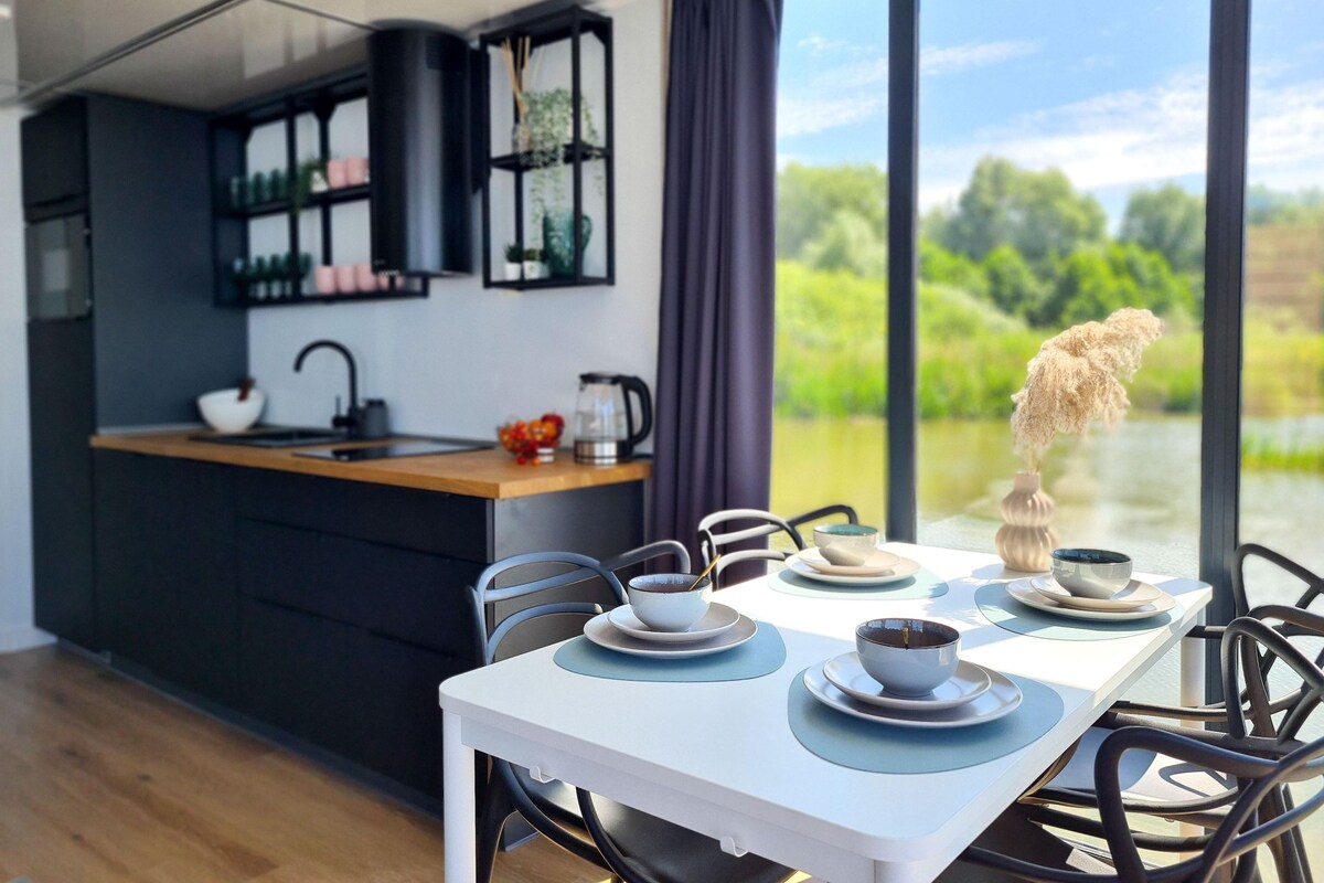 A modern kitchen with a central island, a dining table with chairs, and a view of greenery through large windows.