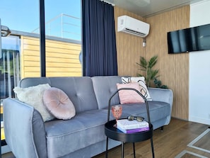 A modern living room with a grey sofa, a small black side table, a mounted air conditioner, a TV, and a large window with curtains. - Luxury Houseboat for 2 People, by the sea (gmina Sianów)